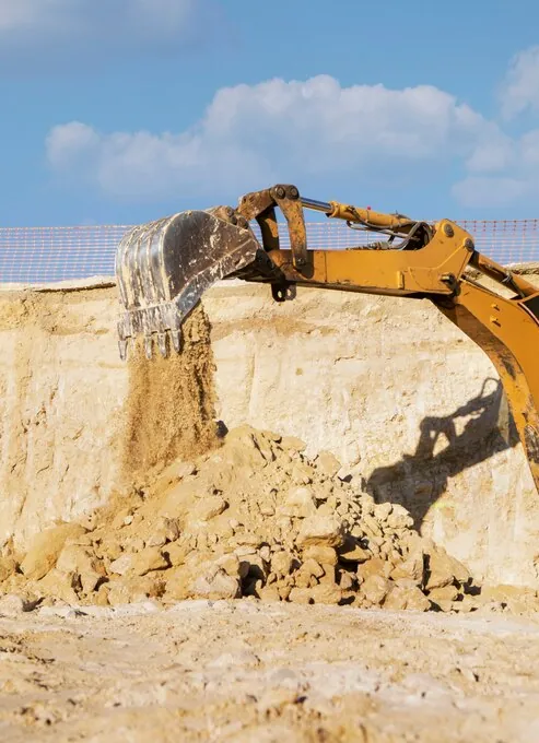 Cupa unui excavator varsă pământ nisipos lângă peretele unei gropi adânci, sub cer senin